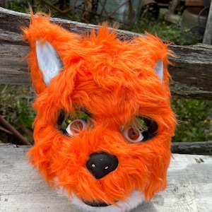 Op de afbeelding: Een feloranje vossenmasker met witte en grijze accenten. Het masker heeft grote ooggaten, een zwarte neus en een pluizige vacht. De oren zijn wit met een grijze binnenkant.