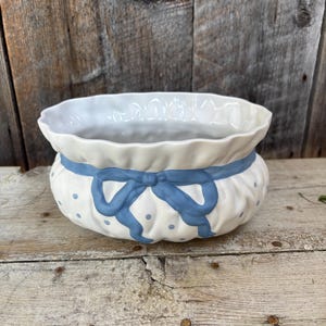 May include: A white ceramic bowl with a ruffled rim and a blue bow design. The bowl has a rounded shape with small blue polka dots and a blue bow tied around the middle. The bowl is sitting on a wooden surface.