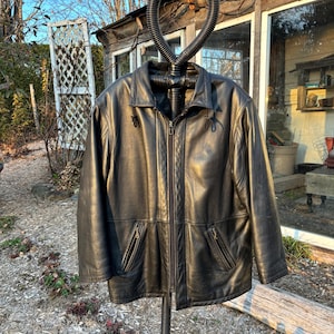 Puede incluir: Una chaqueta de cuero negra con cierre de cremallera y dos bolsillos delanteros. La chaqueta está colgada de un expositor de metal negro. La chaqueta tiene un diseño clásico y un acabado brillante.