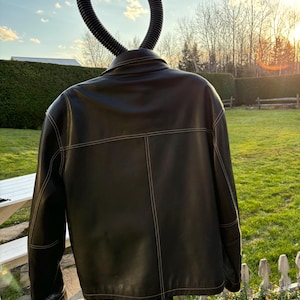 May include: Black leather jacket with white stitching. The jacket is hanging on a hanger against a backdrop of a green lawn and a wooden fence.