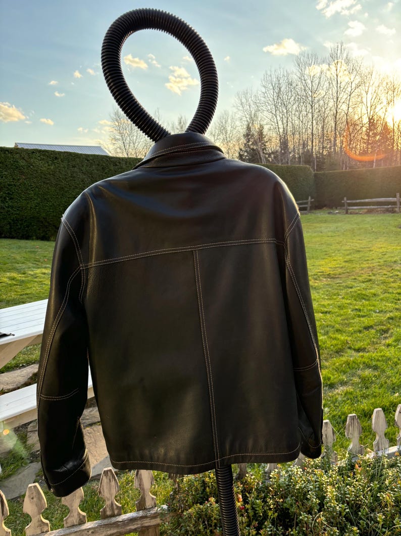 May include: Black leather jacket with white stitching. The jacket is hanging on a hanger against a backdrop of a green lawn and a wooden fence.