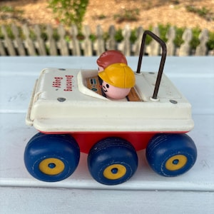 Puede incluir: Un coche de juguete vintage con tres ruedas azules y centros amarillos. El coche tiene una parte superior blanca y una base roja, con dos figuras de juguete en el interior. Las palabras "Bouncing Buggy" están impresas en el lateral.