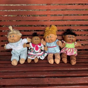 Peut inclure: Quatre poupées Cabbage Patch Kids vintage assises sur un banc en bois. Les poupées ont différentes couleurs de peau et coiffures, portant diverses tenues aux couleurs pastel. Un objet de collection nostalgique.