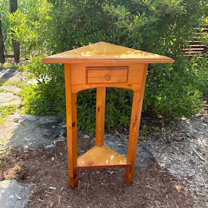May include: A light brown wooden corner table featuring a triangular top, a single centred drawer with a small round knob, and three straight legs connected by a lower triangular shelf. The piece has a smooth, polished finish throughout.
