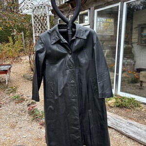 May include: A long black leather coat with a collar and button closure. The coat is hanging on a black stand.
