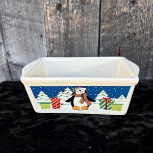 Puede incluir: Molde para pan de cerámica blanca con un diseño de escena de invierno. El diseño presenta un pingüino con un gorro rojo y una bufanda, de pie frente a un bosque nevado con regalos envueltos en verde y rojo.