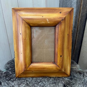 Puede incluir: Un marco de fotos rectangular de madera con un acabado marrón dorado cálido. El marco tiene un borde grueso y biselado y una veta de madera visible. La abertura interior es rectangular, con un respaldo de cartón visible.