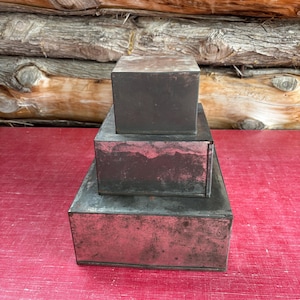 Puede incluir: Tres cajas metálicas rectangulares apiladas con un aspecto envejecido y desgastado. Las cajas son de color gris oscuro con matices rojizos, sobre una superficie roja. El fondo presenta una pared de madera rústica.