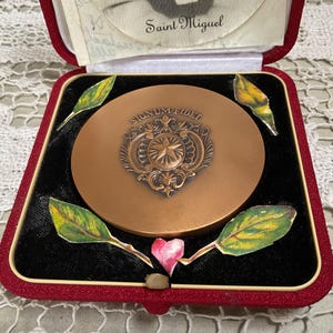 May include: A vintage bronze-colored compact mirror with ornate detailing and the inscription "SIGNUM FIDEL". The compact is in a red velvet-lined box, decorated with painted leaves and a pink heart. The box is open, revealing the mirror.
