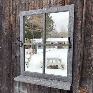 May include: A grey, rectangular window mirror with a distressed, crackled finish. The mirror has a shelf and decorative black metal hardware. The reflection shows a snowy outdoor scene with a picnic table and trees.