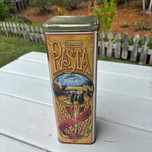 Könnte beinhalten: Rechteckige Pasta-Blechdose mit goldenem und rotem Design. Die Dose trägt das Wort "Lancia Pasta" in einer dekorativen Schriftart. Auf der Vorderseite befindet sich eine Illustration eines Weizenfeldes und von Bauern. Die Worte "Qualité de 1930" stehen auf einem roten Banner.