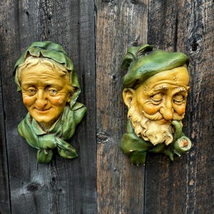 May include: Two vintage wall-mounted sculptures of faces. The sculptures are made of painted plaster and feature a man and a woman with green hats. The man is holding a pipe in his mouth.