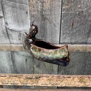 Op de afbeelding: Een bruine keramische plantenbak in de vorm van een kano met een boegbeeld. De plantenbak heeft een glanzende afwerking met groene en gouden accenten. Het boegbeeld heeft groene ogen. De plantenbak wordt gepresenteerd tegen een verweerde houten achtergrond.