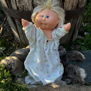Könnte beinhalten: Eine Cabbage Patch Kid Puppe mit blonden Haaren und blauen Augen, die einen weißen und grünen gestreiften Schlafanzug trägt. Die Puppe steht auf einem Stein mit einem braunen und grauen pelzigen Tier daneben.