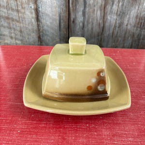 May include: A vintage butter dish with a square lid and base. The dish is a pale yellow-green color with a brown band around the base of the lid. The lid has a small square handle. The dish is sitting on a red surface.