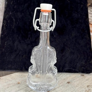 May include: A clear glass bottle in the shape of a violin, with a decorative design. It has a white metal clasp and a white and orange stopper. The bottle is on a wooden surface against a dark background.