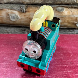 Puede incluir: Un tren de juguete turquesa con una cara sonriente, una chimenea negra y bocanadas de humo amarillas. El tren tiene detalles en rojo y azul. El juguete está sobre un fondo de madera.