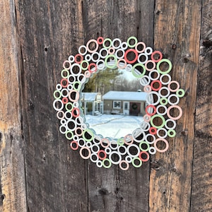May include: A round mirror with a decorative frame made of white, green, and coral-colored circles. The mirror is mounted on a weathered wooden wall, reflecting a snowy outdoor scene.