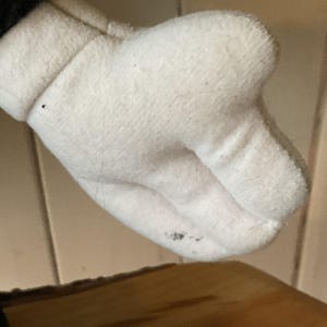 May include: Close-up of a white plush glove with a black cuff. The glove has a rounded shape with visible stitching and a slightly dirty spot on the palm. The background is a light wooden surface.