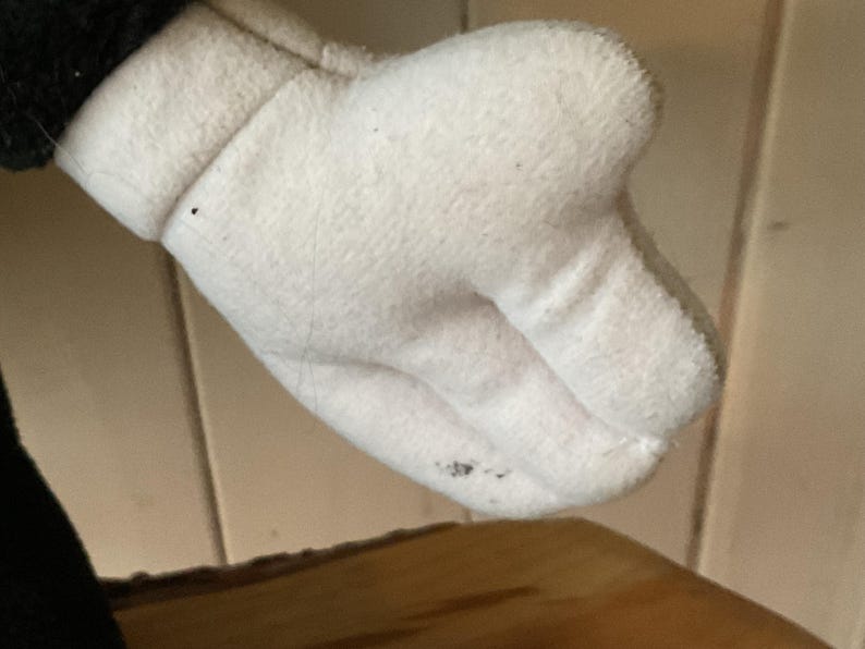 May include: Close-up of a white plush glove with a black cuff. The glove has a rounded shape with visible stitching and a slightly dirty spot on the palm. The background is a light wooden surface.