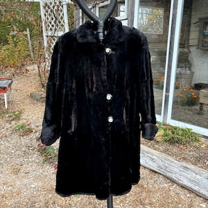 May include: A black faux fur coat with a button closure and long sleeves. The coat is on a black stand in front of a wooden building.