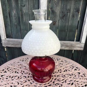 May include: A classic oil lamp with a deep red base and a white, textured shade. It has a clear glass chimney and rests on a light pink, ornate table. The background features a weathered wooden fence.