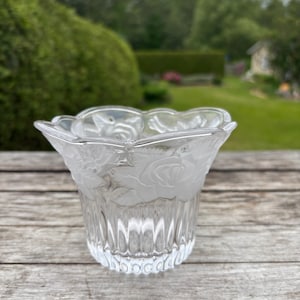 Peut inclure: Un photophore en verre transparent avec un motif floral givré. Le bord supérieur a une forme festonnée et le corps présente des rainures verticales. Le motif givré représente des roses. Le photophore est posé sur une surface en bois.