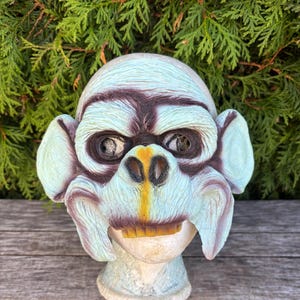 May include: A light blue and brown monkey mask with large ears, a wide mouth, and visible teeth. The mask has a textured surface and is mounted on a light blue stand. The mask is likely for Halloween or costume use.
