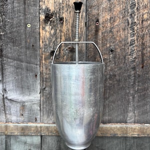 May include: A vintage, cylindrical aluminum butter churn with a black handle and a spring mechanism. The churn has a rounded base and a metal handle. The background is a weathered wooden surface.