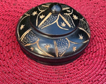 Può includere: Una scatola rotonda di legno nero con coperchio. La scatola e il coperchio sono decorati con motivi intagliati a stella e floreali in un colore più chiaro. La scatola è su una superficie rossa intrecciata.