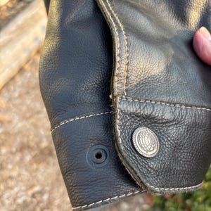 May include: Close-up of a black leather jacket cuff with a button and stitching detail. The stitching is a light brown color.