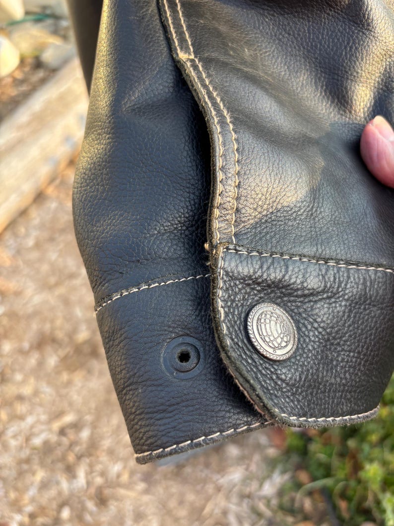 May include: Close-up of a black leather jacket cuff with a button and stitching detail. The stitching is a light brown color.