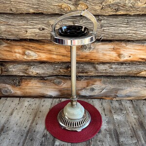 Puede incluir: Un cenicero vintage con un cuenco negro y un soporte de metal plateado. El cenicero tiene un asa curvada y se apoya sobre una alfombra roja tejida. La base tiene un diseño decorativo y un elemento de piedra verde claro.