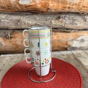 May include: A set of four white ceramic mugs stacked in a chrome metal rack. Each mug features a different festive design: colorful stripes, Christmas trees, snowflakes, and ornaments. The mugs sit on a red woven placemat, with a wooden log background.