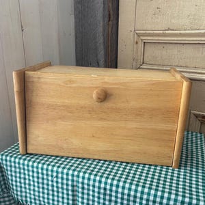 May include: A light-coloured wooden bread bin with a rounded knob on the front. The box is rectangular with a lid and sits on a green and white checked tablecloth. The wood grain is visible.