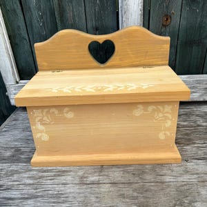 May include: A wooden box with a heart-shaped cutout in the lid. The box is painted a light brown colour and has a white floral design painted on the front. The box is sitting on a wooden surface.