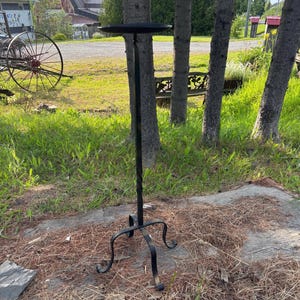 May include: A tall, black wrought iron plant stand with a circular top and a twisted stem. The stand has three curved feet and is set outdoors on a bed of pine needles and grass.