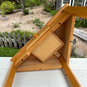 May include: A light brown wooden desk organiser featuring an angled top surface with a small drawer and a central knob. The structure includes a flat base, side supports, and a rectangular compartment with a light-coloured panel, showing visible screw holes.