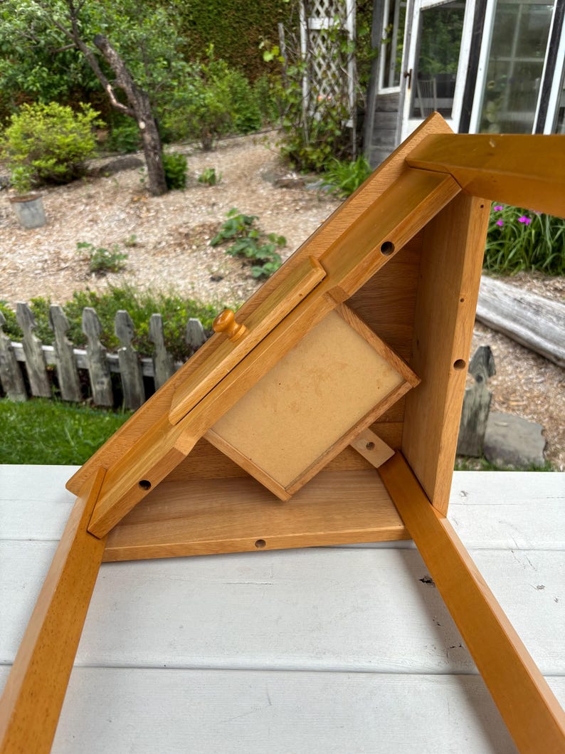May include: A light brown wooden desk organiser featuring an angled top surface with a small drawer and a central knob. The structure includes a flat base, side supports, and a rectangular compartment with a light-coloured panel, showing visible screw holes.