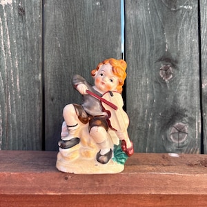 May include: A vintage ceramic figurine of a child playing a violin. The figurine features a seated child with red hair, wearing a gray jacket, brown shorts, and black shoes. The violin is cream-colored with a red bow. The figurine is on a cream-colored base.