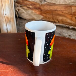May include: A white ceramic mug featuring a black background with a graphic print of a green character with orange hair. The mug has a white handle and a white interior, showcasing a cylindrical shape with a smooth, glossy finish throughout the exterior.