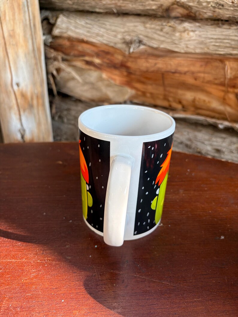 May include: A white ceramic mug featuring a black background with a graphic print of a green character with orange hair. The mug has a white handle and a white interior, showcasing a cylindrical shape with a smooth, glossy finish throughout the exterior.