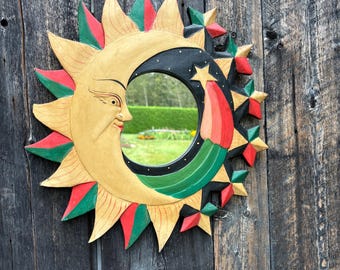 May include: A wooden sun and moon mirror with a smiling face, surrounded by star accents. The sun is painted yellow with red and green accents. The moon is black with a star and a red and yellow streak. The mirror is round.