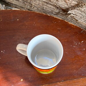 May include: A ceramic mug with a white interior and a handle on the left side. The exterior features a horizontal band of bright orange and lime green colors. The mug is viewed from a top-down angle, showing the circular opening and the smooth rim.