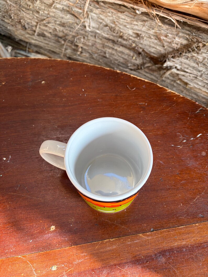 May include: A ceramic mug with a white interior and a handle on the left side. The exterior features a horizontal band of bright orange and lime green colors. The mug is viewed from a top-down angle, showing the circular opening and the smooth rim.