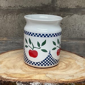 Pode incluir: Jarro de cerâmica branca com um padrão xadrez azul e branco e maçãs vermelhas com folhas verdes.