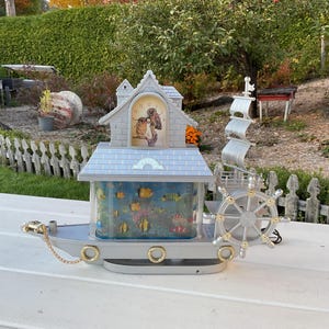 May include: A grey and white decorative clock shaped like a boat with a small aquarium and a clock face featuring a picture of an owl. The clock has a gold chain and a gold steering wheel.