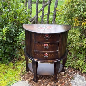 Peut inclure: Une commode demi-lune en bois foncé dotée de trois tiroirs superposés avec des motifs floraux peints à la main et des poignées en métal. Le plateau présente un motif floral peint et l'unité repose sur quatre pieds incurvés.