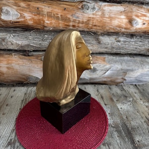 Op de afbeelding: Een goudkleurige buste van een vrouw met gesloten ogen en vloeiend haar. De sculptuur staat op een zwarte rechthoekige basis, geplaatst op een rode cirkelvormige mat. De achtergrond bestaat uit houten stammen.
