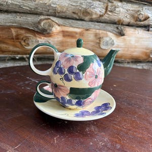 Peut inclure: Un service à thé en céramique pour une personne composé d'une théière empilée sur une tasse et une soucoupe. L'ensemble présente une base crème ornée de grappes de raisins violets, de feuilles vertes et de fleurs roses peintes à la main.